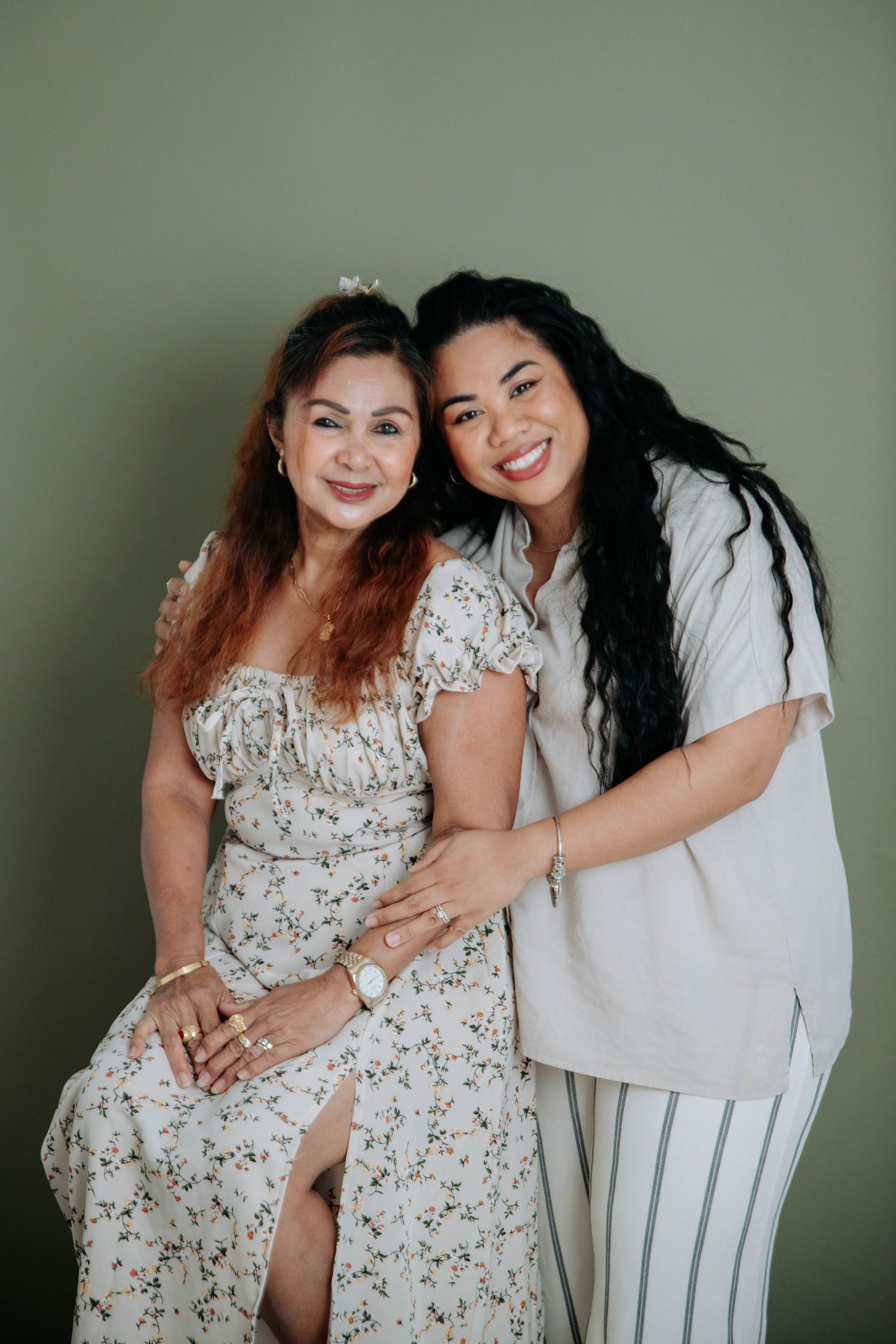 Two people in floral and neutral outfits pose together against a sage green background.