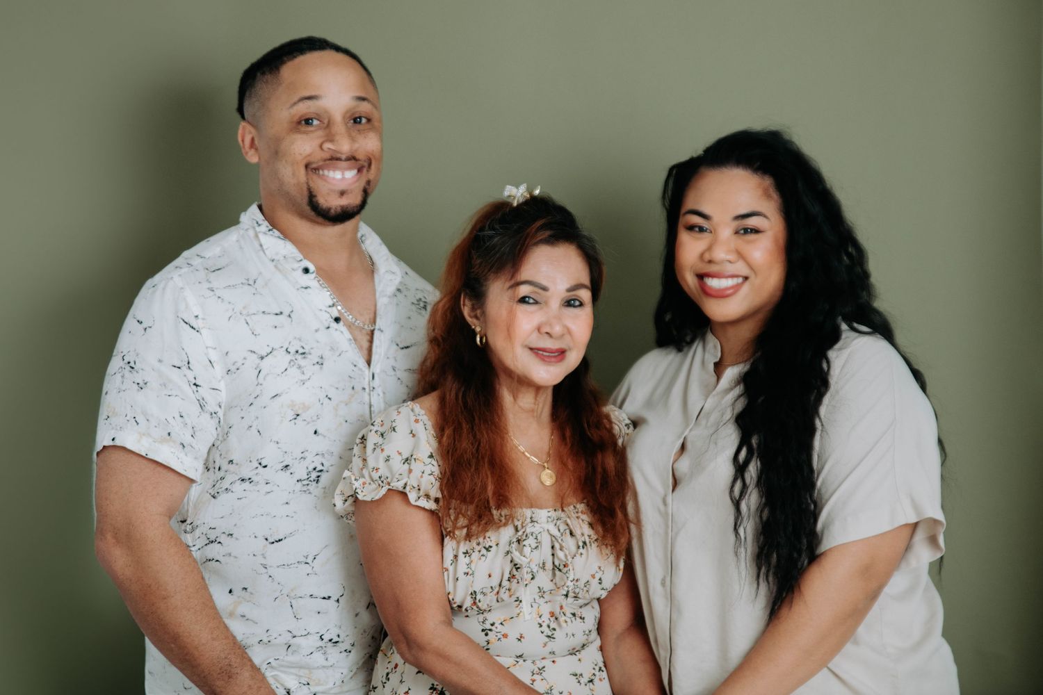 Three people in coordinated outfits pose together against a sage green background.