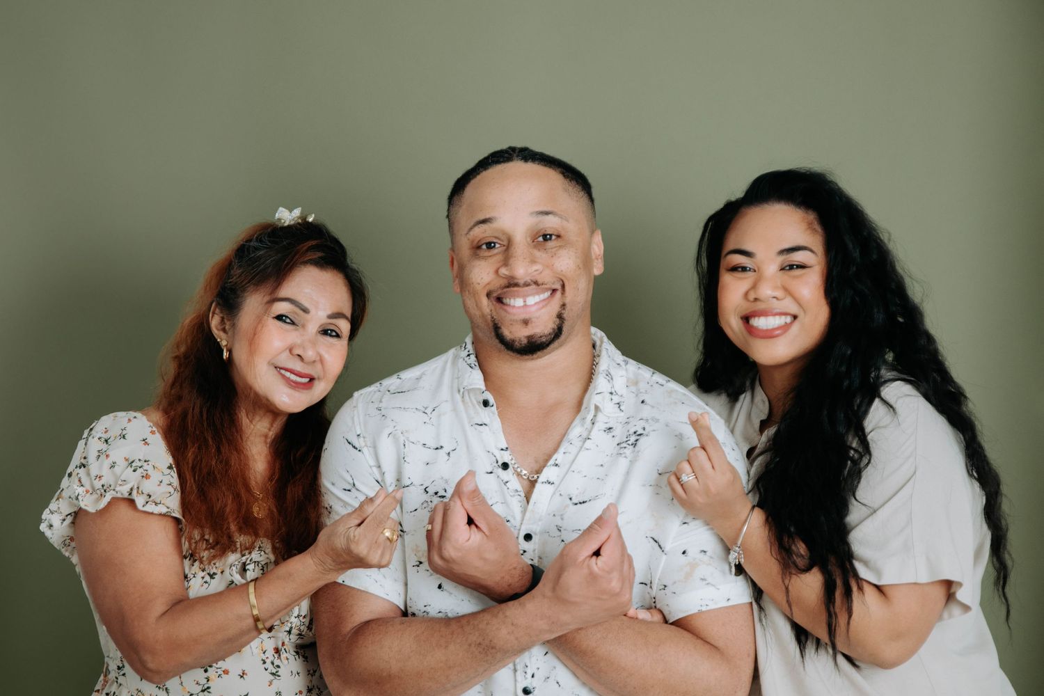 Three people pose together wearing white and cream colored outfits against a green studio backdrop.