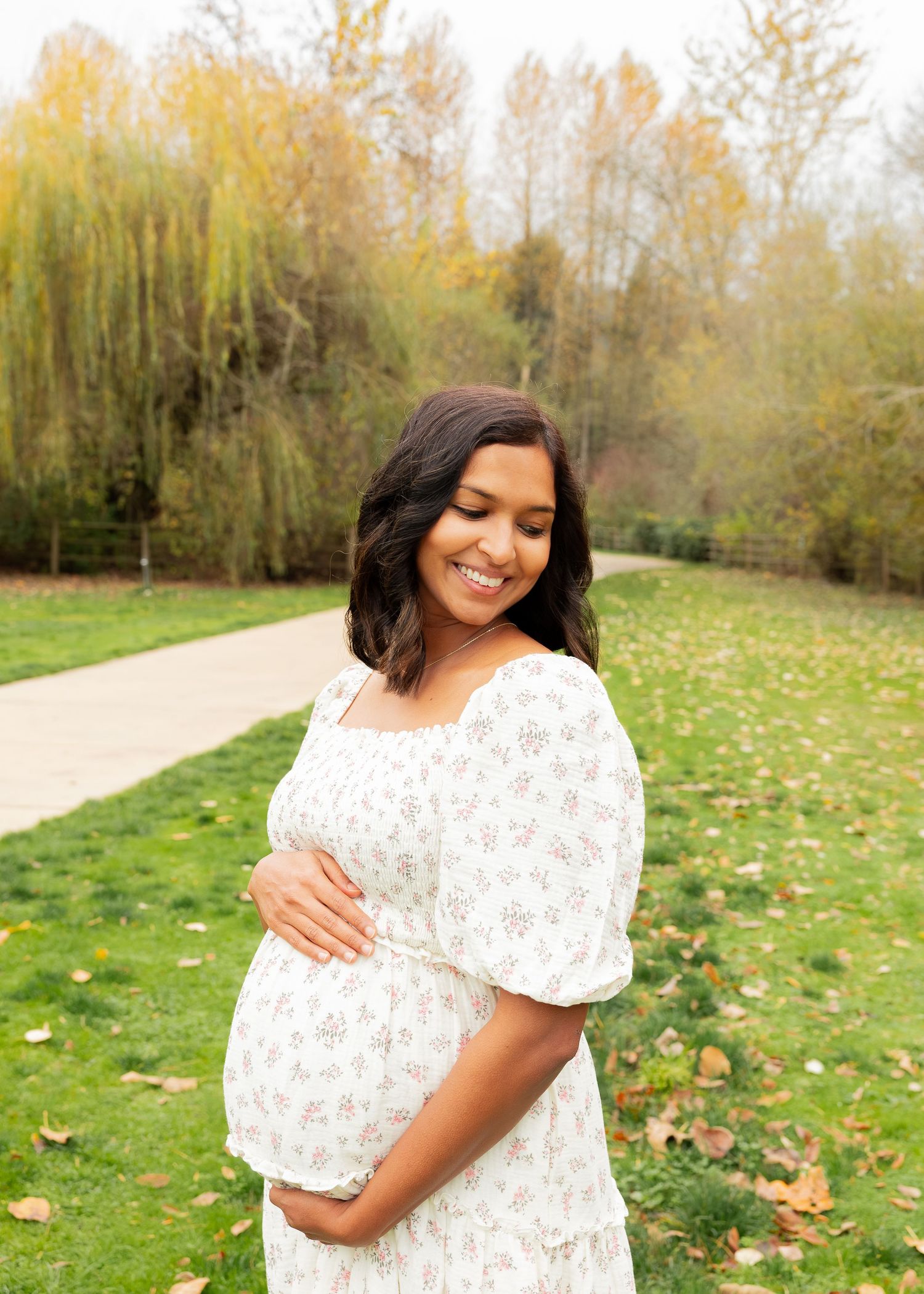 Expectant mother in white dress smiling while cradling baby bump in autumn park.