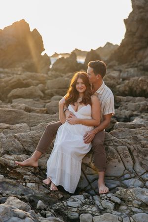 Romantic couple shares tender moments on rocky coastal shore during golden hour sunset photoshoot.