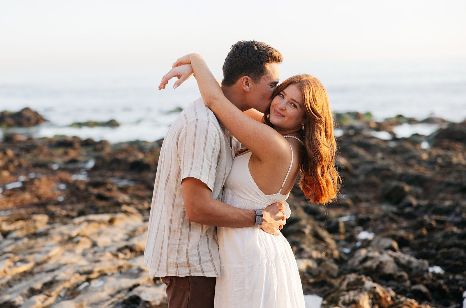 A couple embraces on a rocky beach during sunset with golden light illuminating their silhouettes.