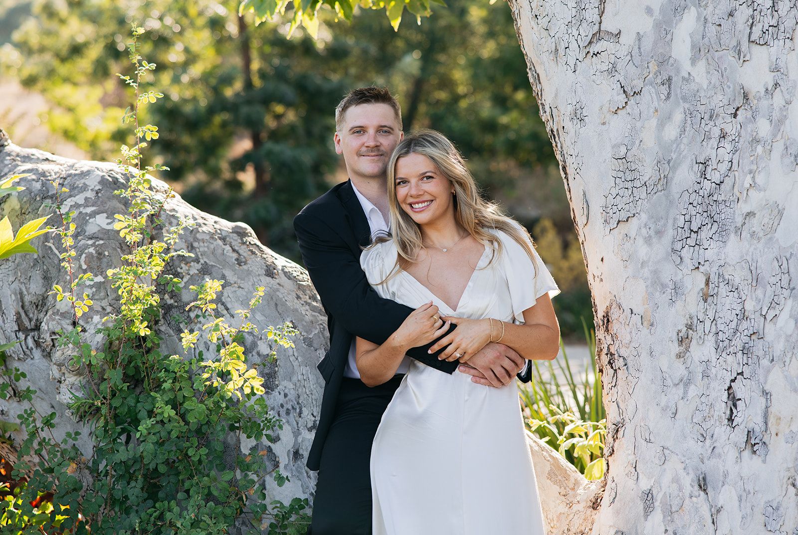 Happy couple embraces and laughs together in sunlit garden setting with white tree trunks behind them.