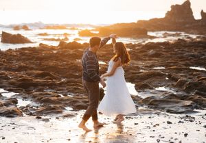 Couple walking hand in hand along rocky beach shore during golden sunset with ocean waves crashing.