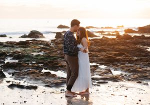 Romantic couple embraces on rocky beach at sunset with ocean waves crashing in background.
