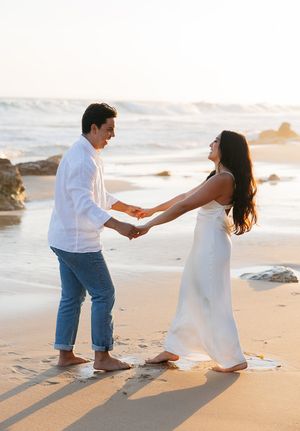 A couple holding hands and dancing together at sunset on a beach with waves and rocks in the background.