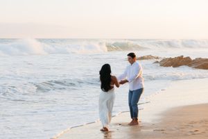 Two people holding hands on a beach at sunset while ocean waves crash against rocks in the background.