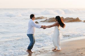 A couple holds hands and dances together on a beautiful beach at sunset with ocean waves in the background.