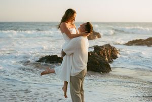A romantic couple embraces on a beach at sunset with waves crashing against rocks in the background.