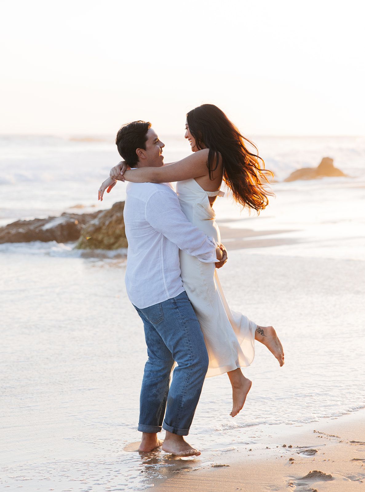 A romantic couple in white and denim embraces while standing barefoot on a beach at sunset by the ocean.