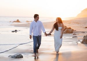 A couple walking hand in hand along a scenic beach at sunset with waves and rocks in the background.