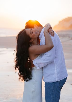 A romantic couple embracing on a beach during golden hour with sunlight creating a warm glow behind them.