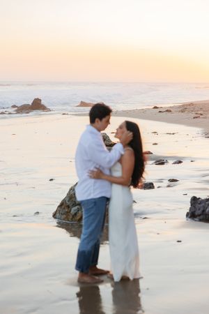 A romantic couple embraces on a scenic beach at sunset with gentle waves and rocky outcrops in the background.