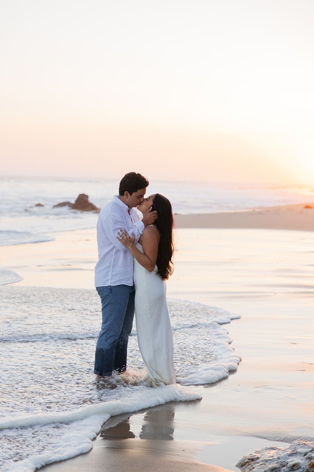 A couple embraces romantically on a sunny beach at sunset while waves gently lap at the shoreline.