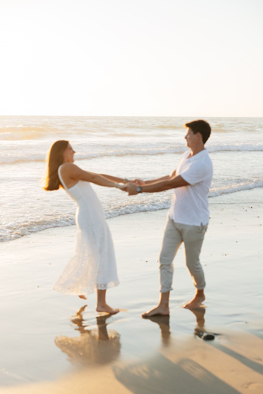 Couple in white and gray holding hands and dancing together on a sandy beach at sunset.