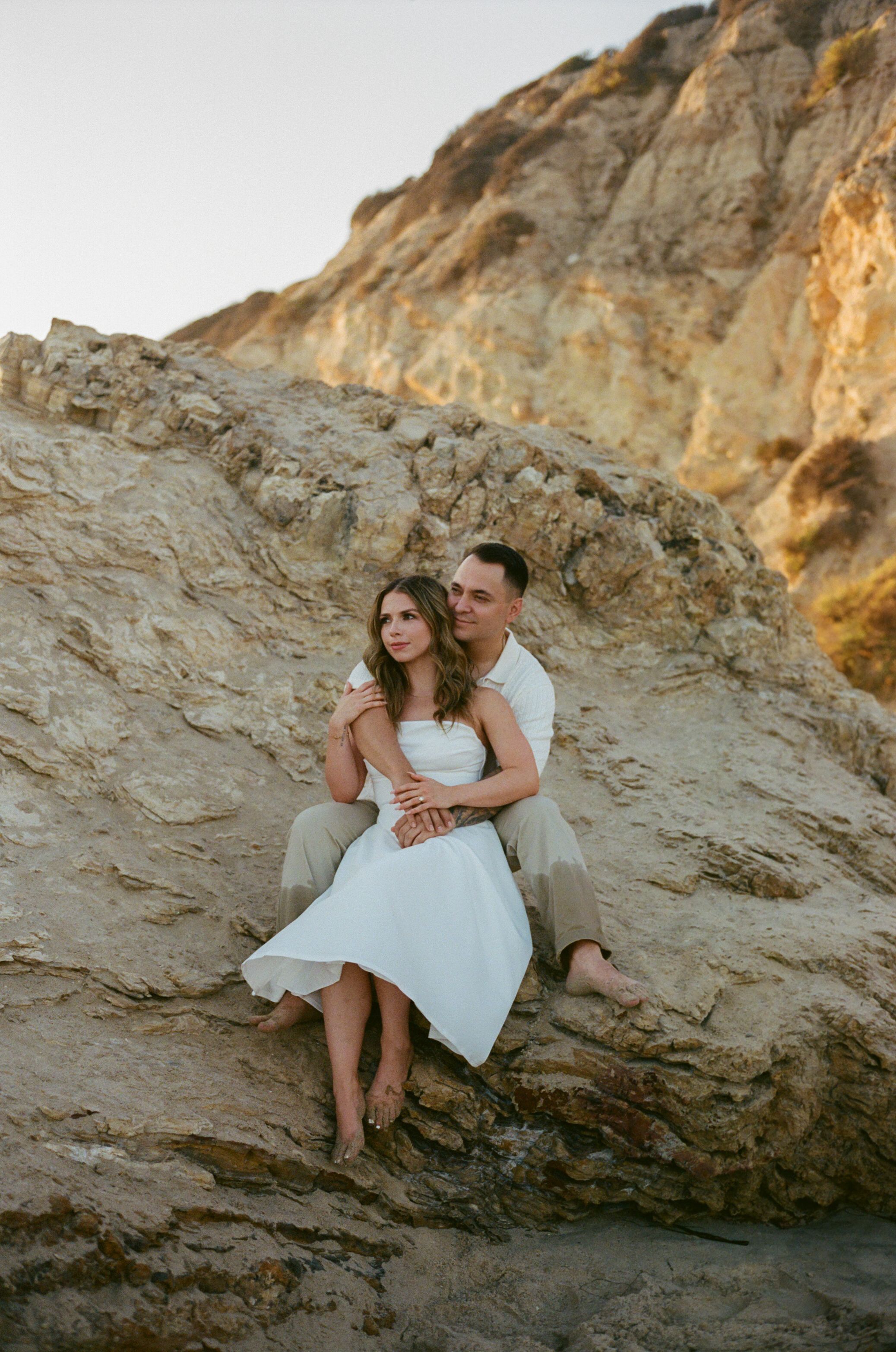 A couple in white outfits sits together on rocky coastal cliffs during golden hour at the beach.