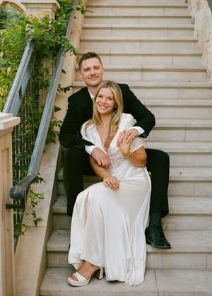A couple in wedding attire sits and poses together on white marble stairs with greenery in the background.