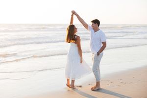 A couple dancing together on a serene beach at sunset, wearing white and light colored outfits.
