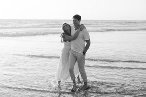 A romantic couple embraces while standing at the edge of the ocean in a black and white beach photo.