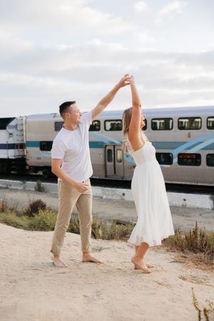 A couple dances playfully near train tracks with a commuter train in the background.