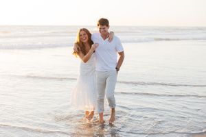 A couple walking together at sunset along the beach shoreline in white and light colored clothing.