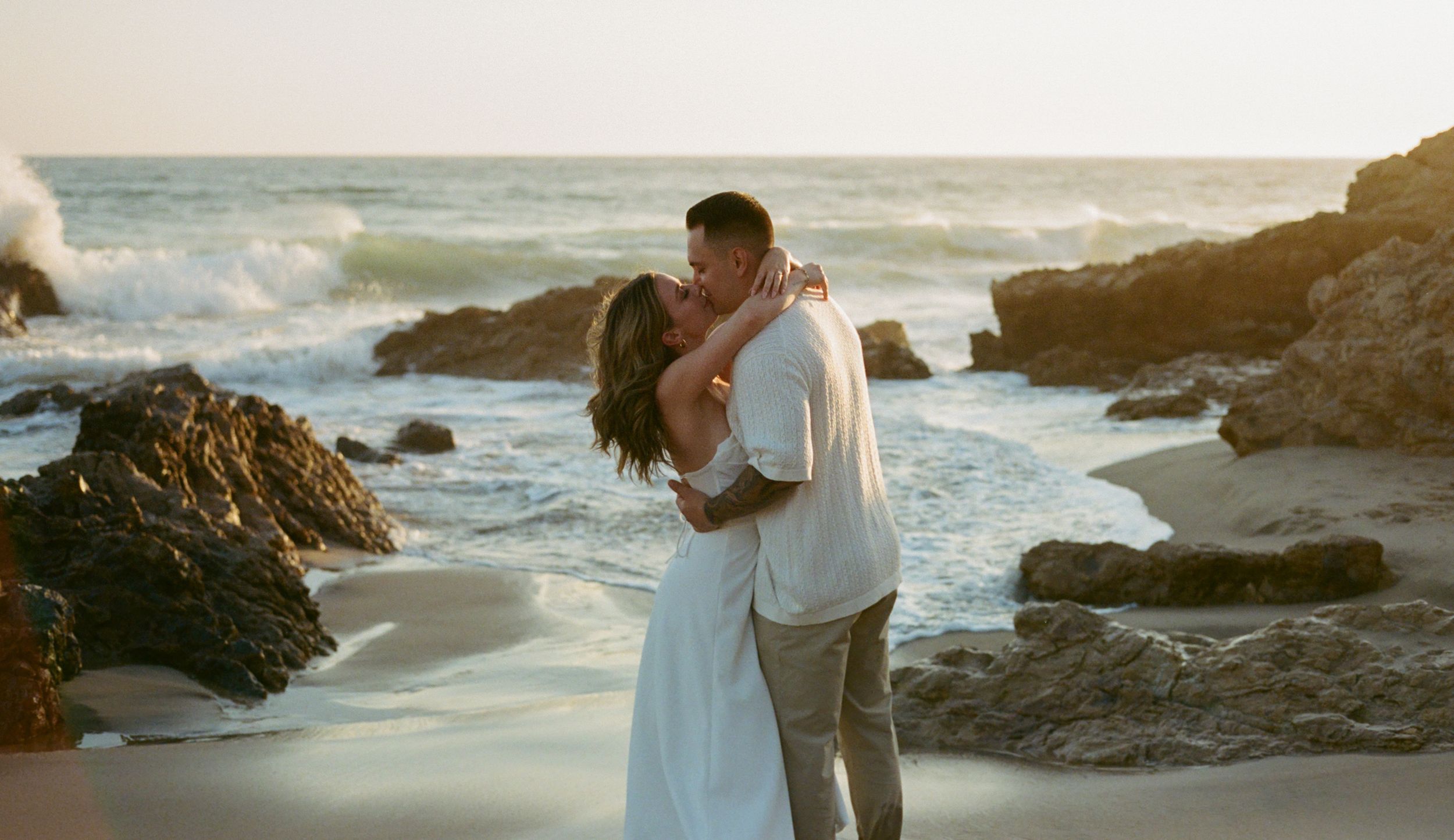 A romantic couple embraces on a rocky beach at sunset with crashing waves and coastal rocks in the background.
