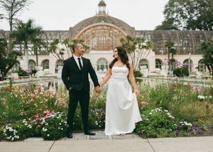 A couple walks through a lush garden with beautiful rose bushes and elegant archways at a stately venue.