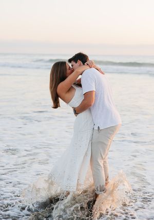 A romantic couple embraces in the ocean waves at sunset on a beach, water splashing around their feet.