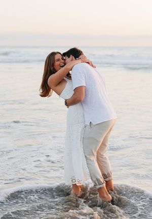 A romantic embrace in the ocean waves at dusk with a couple wearing white and light colored outfits.