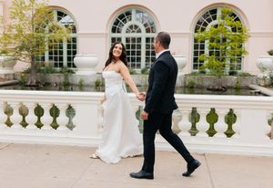 A couple in formal wedding attire walking along an elegant balcony with arched windows and columns.