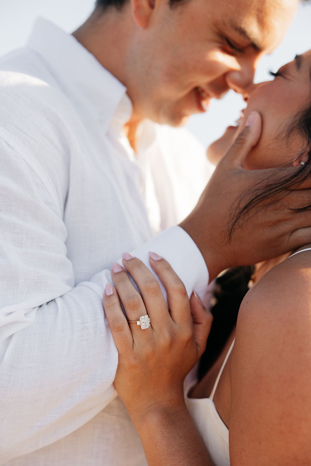 Close up engagement photo showing a sparkling diamond ring while couple shares an intimate moment in white attire outdoors.