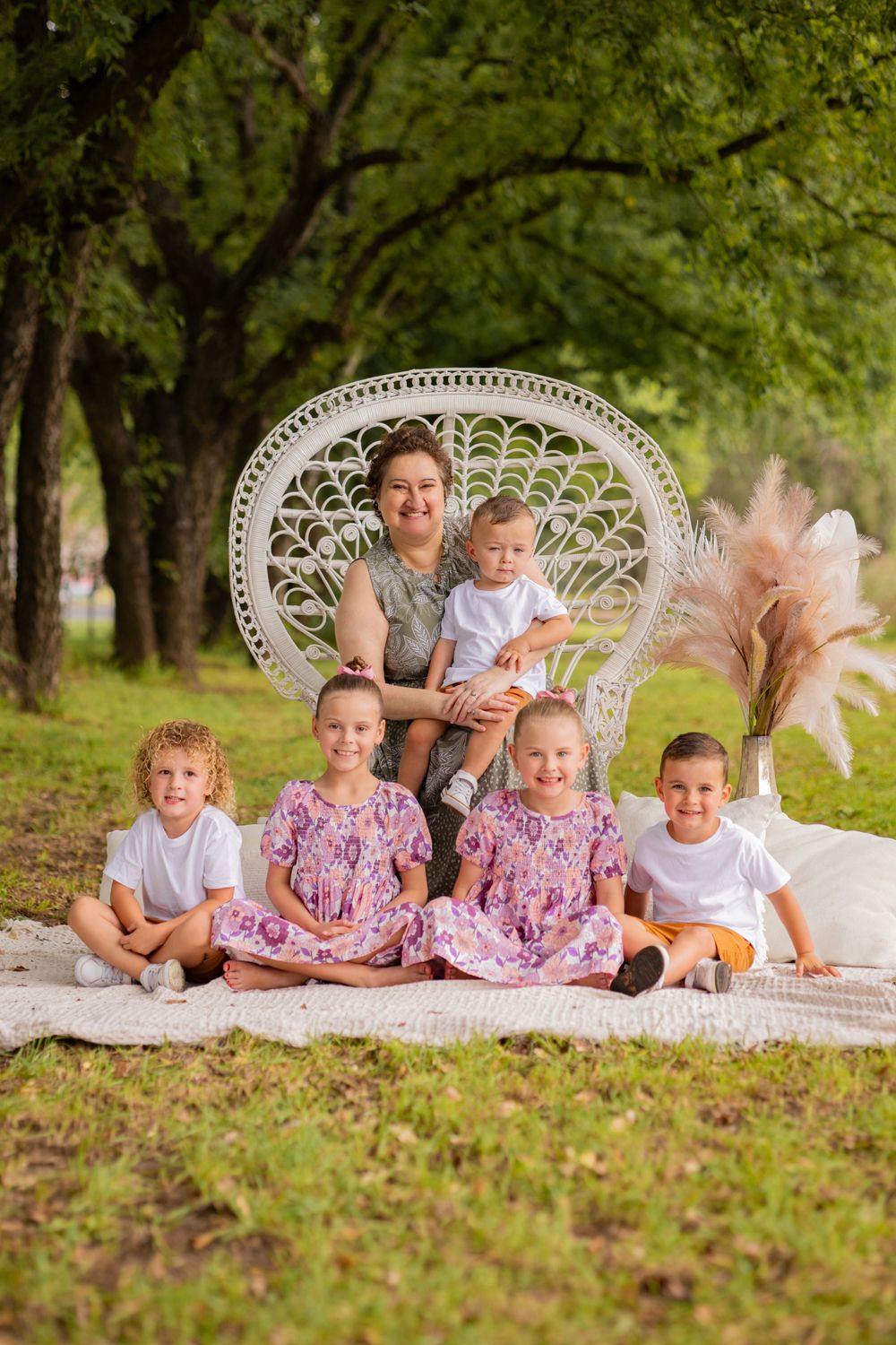 Woman with six young children posing outdoors in park with white peacock chair.