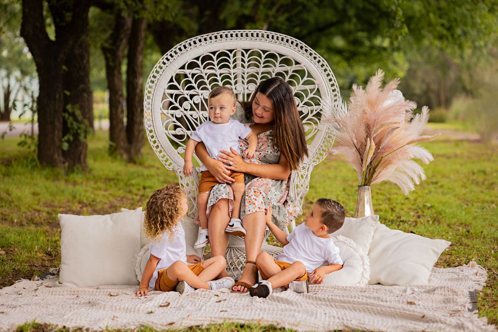 Mother sitting in peacock chair with three young children on outdoor blanket.