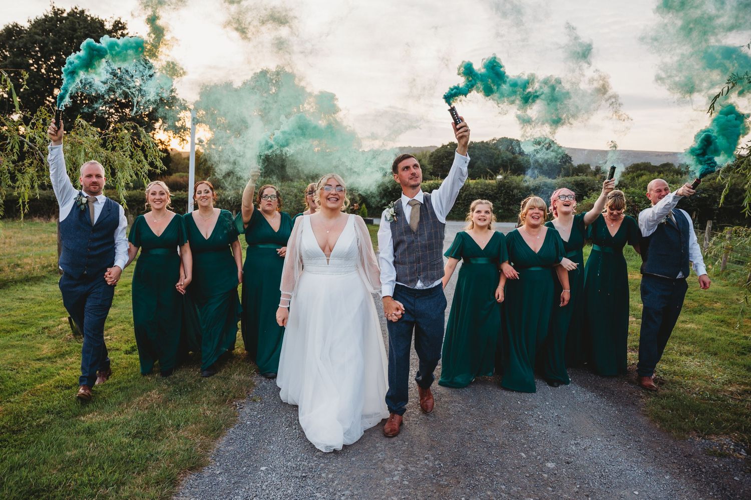 Wedding party celebrates with teal smoke bombs on countryside path.