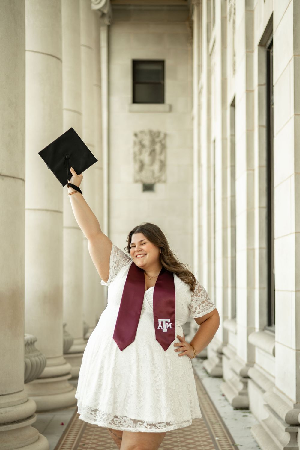 Graduate in white dress and burgundy stole poses joyfully between classic white columns during graduation photoshoot.