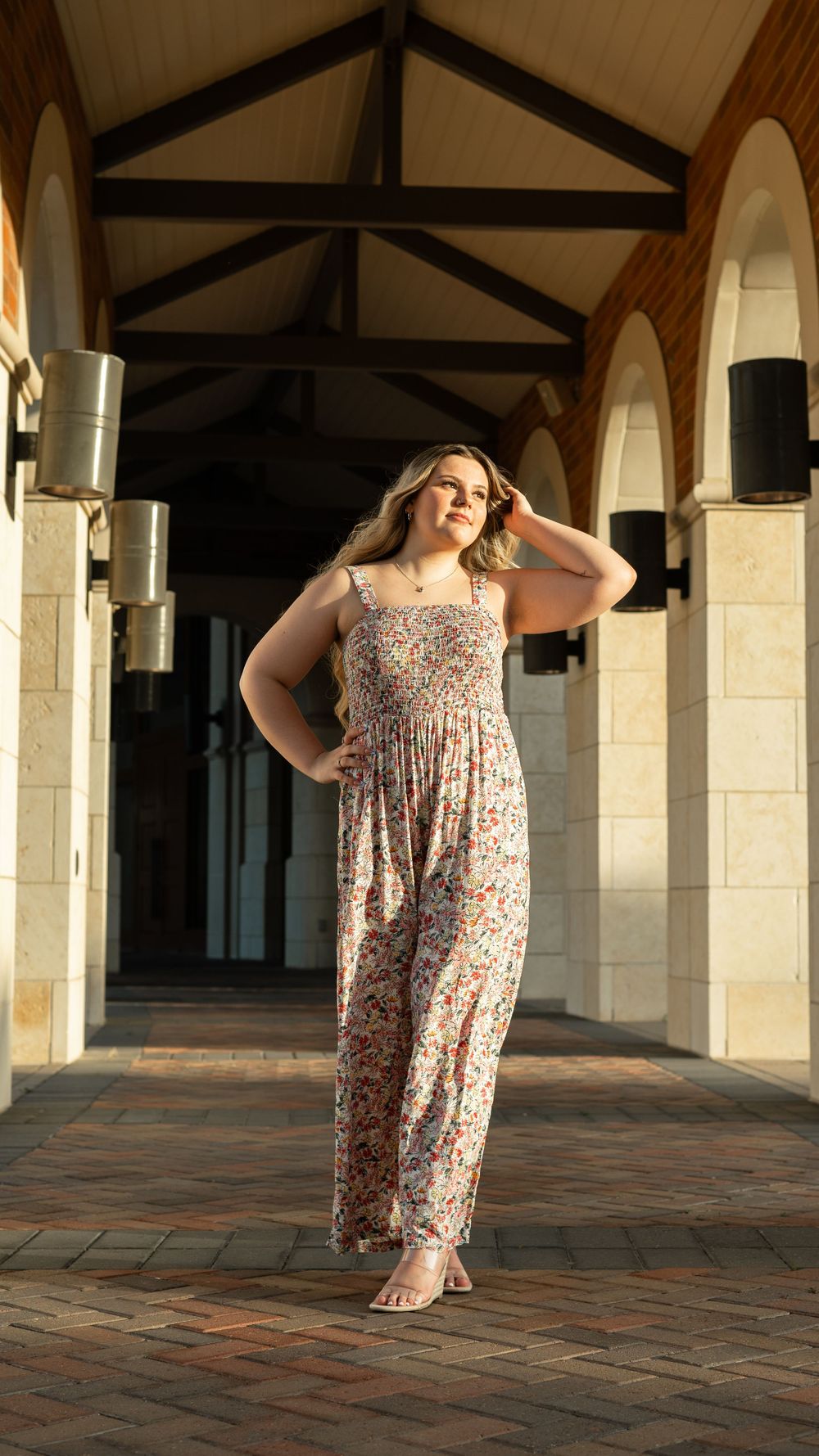 A person poses in a floral maxi dress on a brick patio under arched columns and exposed wooden beams.