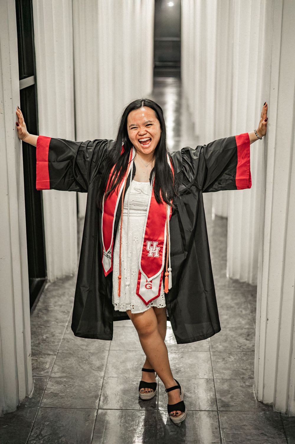 A graduate in black cap and gown poses joyfully between white pillars during a graduation photoshoot.