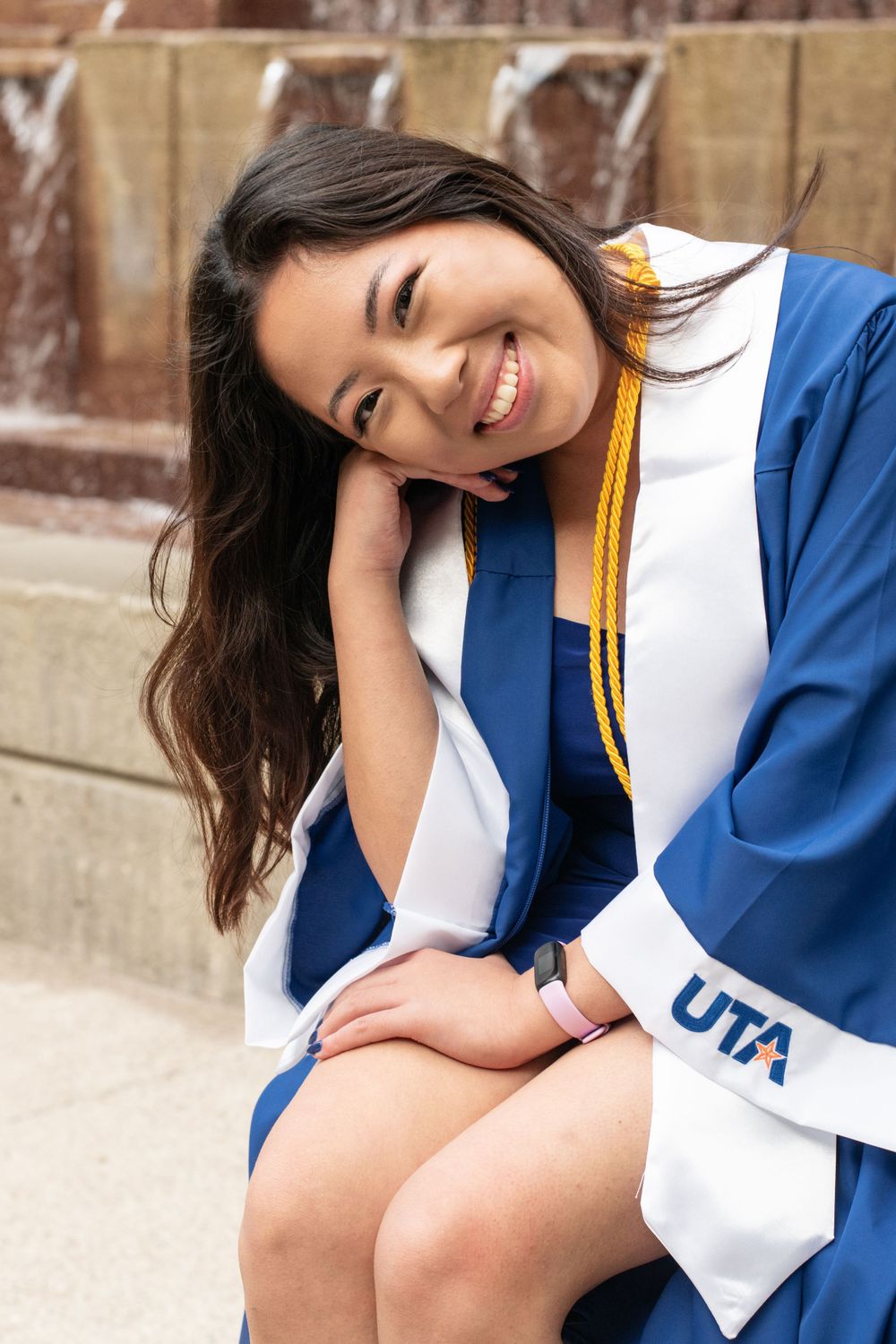 Graduate in blue and white UTA graduation gown smiling while sitting on stone steps.