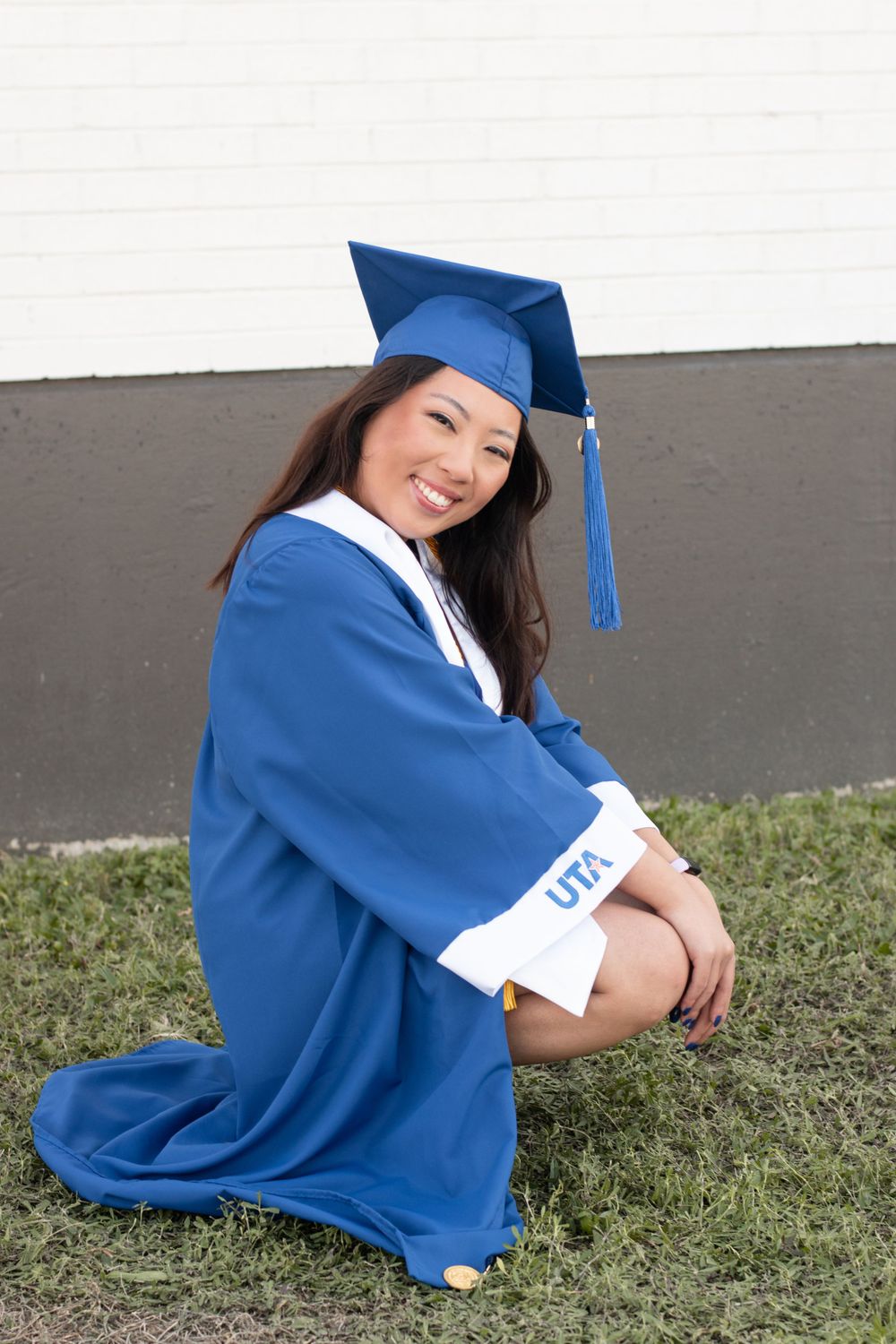 Graduate in blue cap and gown poses seated on grass in front of dark wall.