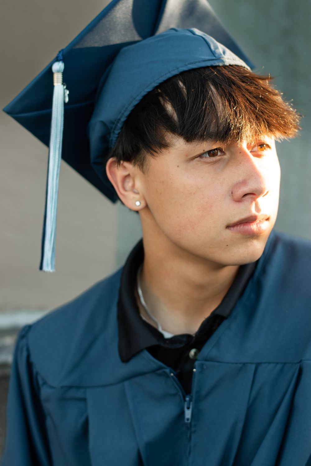A graduate in navy blue cap and gown looks thoughtfully to the side during a graduation photo session.