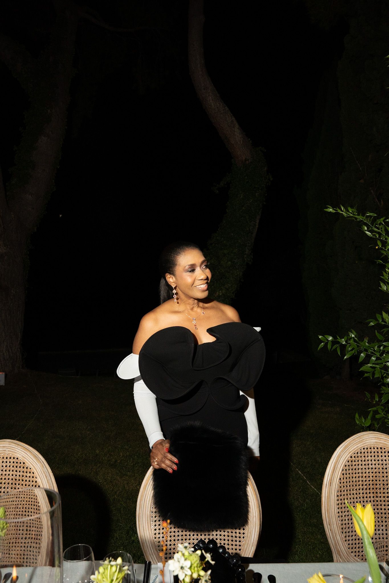 Elegant figure in black off-shoulder dress poses by rattan chairs at outdoor evening dinner table with floral arrangements.