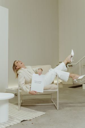 Model in white outfit reclines on modern chair with paper in minimalist setting.