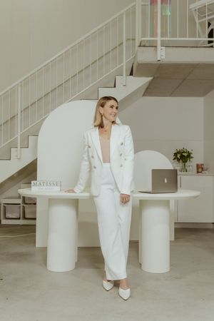 Figure in white power suit poses between cylindrical tables against industrial white interior with stairs.