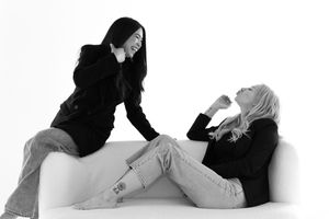 Two people in black clothing pose casually on white background in artistic black and white portrait.