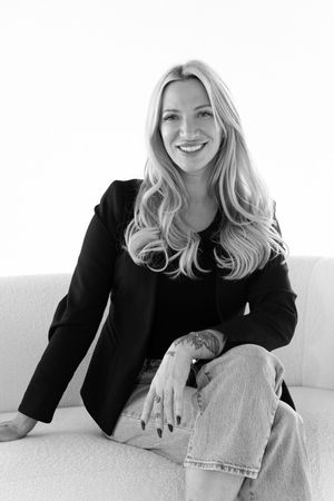 Black and white portrait of someone in a black blazer and light jeans sitting casually on a white couch.