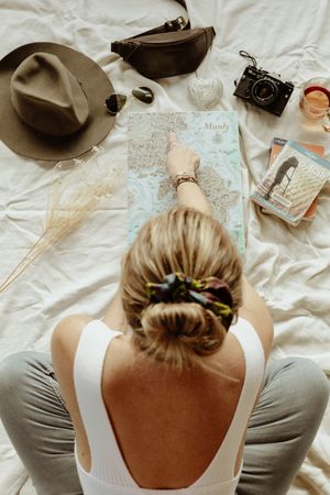 A person with blonde hair in a white tank top studies a map while sitting on a white surface.