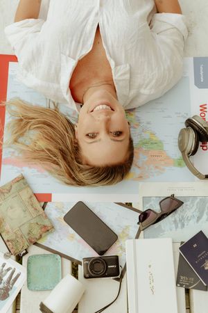 Travel planning flatlay with maps, camera, phone and other items surrounding someone lying on white background.