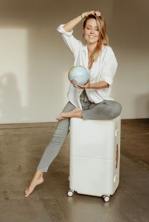 Person in white shirt and gray pants sits on white luggage holding small globe while posing against neutral wall.