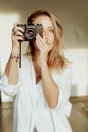 Person in white shirt holding vintage camera against neutral wall with soft lighting.