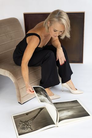 Person in black attire sits on beige chair looking at large photography book with monochrome images.